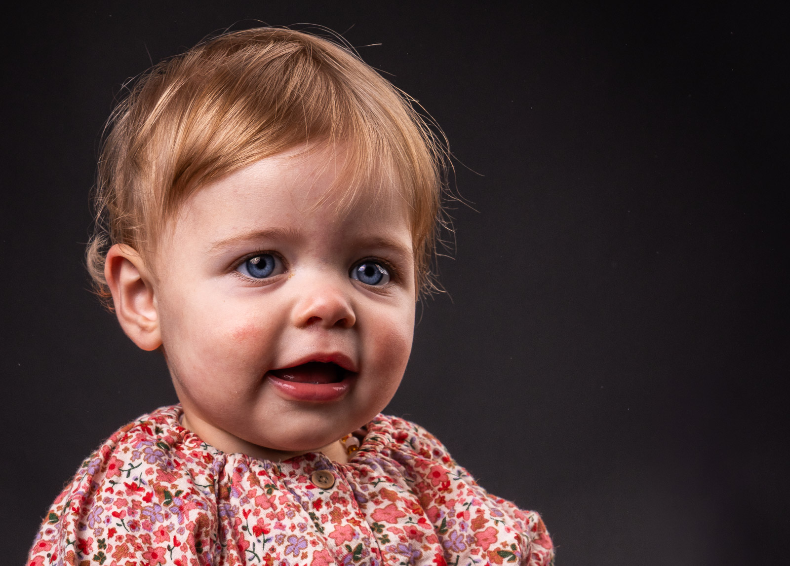 Schattig babyportret van een meisje met blauwe ogen en een gebloemd shirt tegen een donkere studioachtergrond.