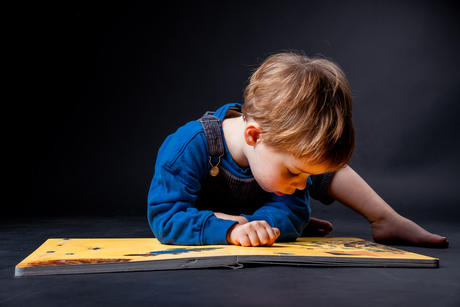 Geconcentreerde peuter liggend op zijn buik die een geel prentenboek leest tijdens een kinderfotografie studioshoot.